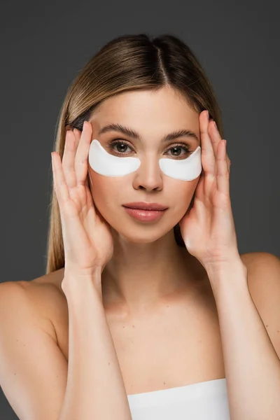 Bonita mujer con parches de colágeno en los ojos posando con las manos cerca de la cara aislada en gris - foto de stock