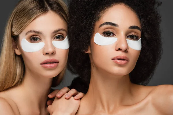 Multicultural women with naked shoulders and eye patches looking at camera isolated on grey — Stock Photo