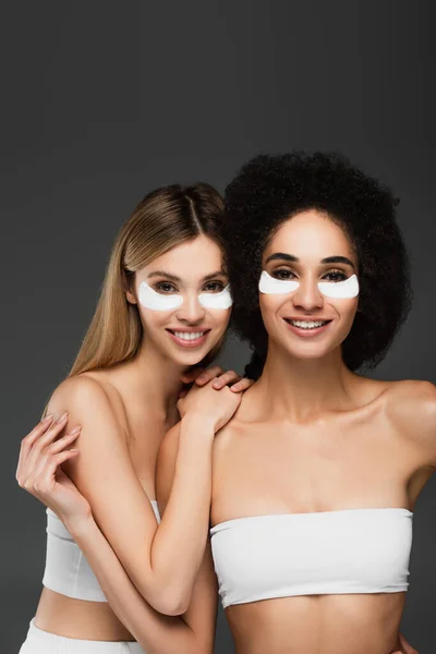 Alegres mujeres multiétnicas en parches de hidrogel sonriendo a la cámara aislada en gris - foto de stock