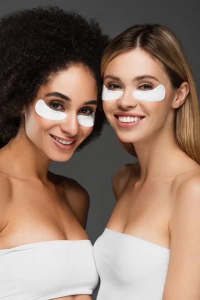 Jeunes femmes interracial dans les yeux patchs souriant à la caméra isolé sur gris — Photo de stock