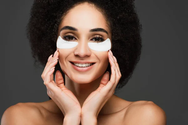 Alegre mujer afroamericana en parches de ojos tocando la cara mientras sonríe a la cámara aislada en gris - foto de stock