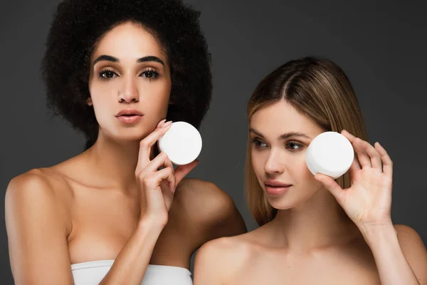 Mujeres multiétnicas con piel perfecta mostrando recipientes con crema cosmética aislada en gris - foto de stock