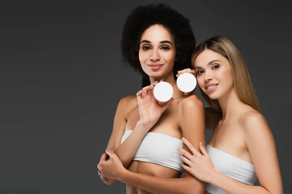 Mulheres multiculturais sorridentes com pele limpa posando com creme corporal isolado em cinza — Fotografia de Stock