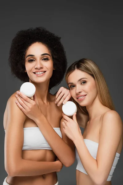 Cheerful interracial women in white tops posing with cosmetic cream isolated on grey — Stock Photo