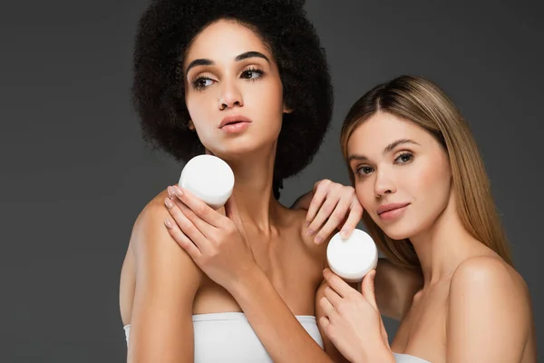 Junge und hübsche multiethnische Frauen mit perfekter Haut und natürlichem Make-up posieren mit kosmetischer Creme isoliert auf grau — Stockfoto