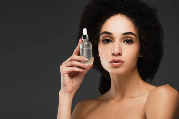 Pretty african american woman with bare shoulders holding bottle of cosmetic serum isolated on grey — Stock Photo