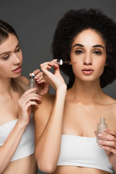 Joven mujer afroamericana mirando a la cámara mientras aplica suero cosmético aislado en gris - foto de stock