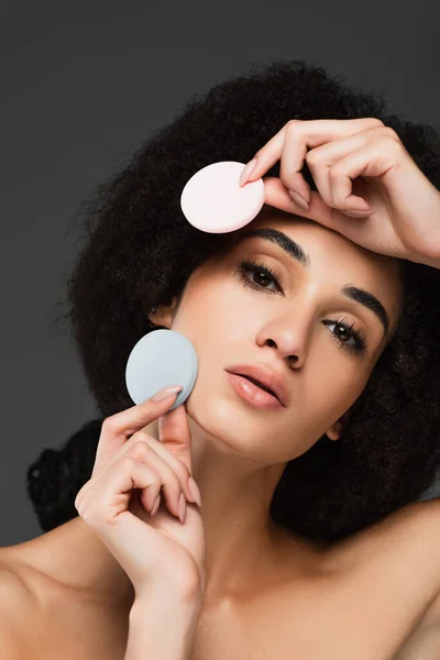 Joven mujer afroamericana con piel perfecta y maquillaje natural posando con esponjas cosméticas aisladas en gris - foto de stock