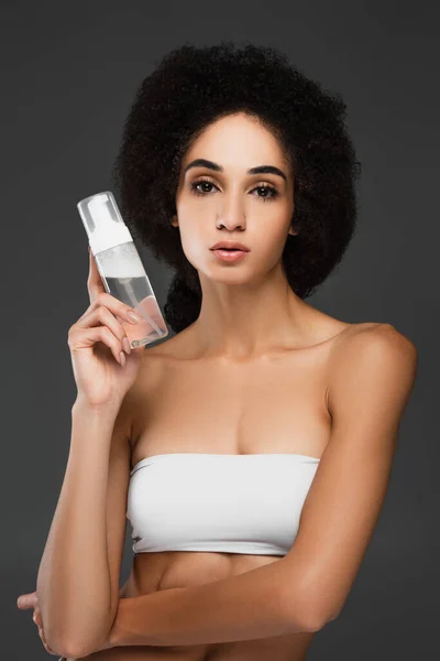 Pretty african american woman in white top looking at camera while holding makeup remover isolated on grey — Stock Photo