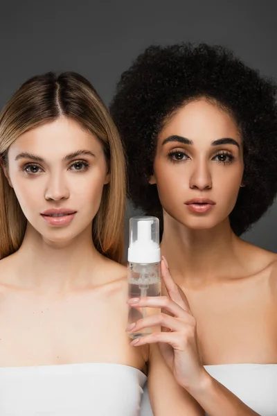 Junge und hübsche multikulturelle Frauen mit sauberer Haut, die in die Kamera schauen, in der Nähe von Kosmetikschaum isoliert auf grau — Stockfoto