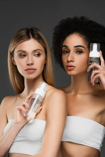 Mujeres multiculturales en tapas blancas sosteniendo botellas con espuma cosmética aislada en gris - foto de stock
