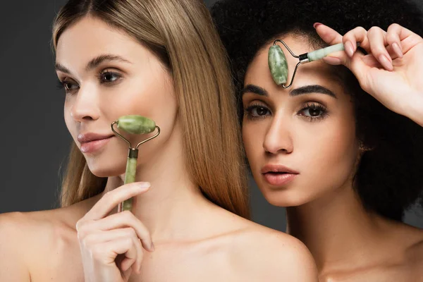 Jovem interracial mulheres massagear rostos com jade rolos isolado no cinza — Fotografia de Stock