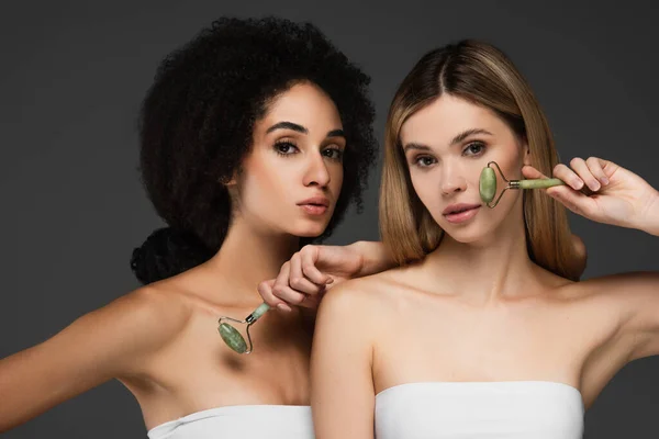 Mulheres interracial sensuais com pele perfeita e maquiagem natural posando com rolos de jade isolados em cinza — Fotografia de Stock