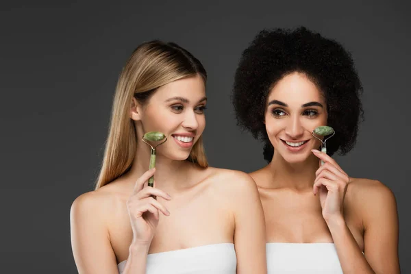 Mulheres multiétnicas felizes fazendo massagem facial com rolos de jade isolados em cinza — Fotografia de Stock