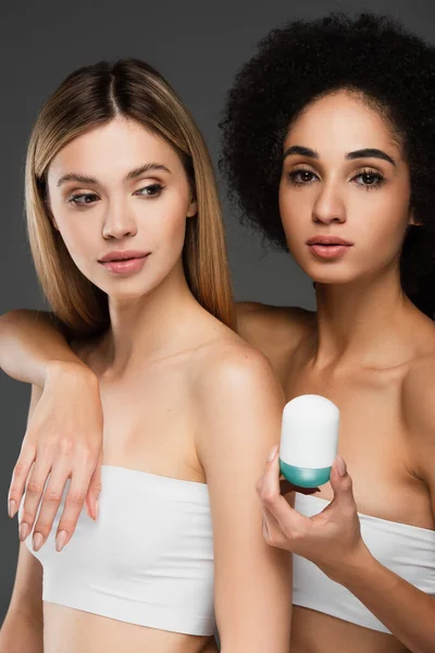 Young african american woman embracing pretty friend while holding deodorant isolated on grey — Stock Photo