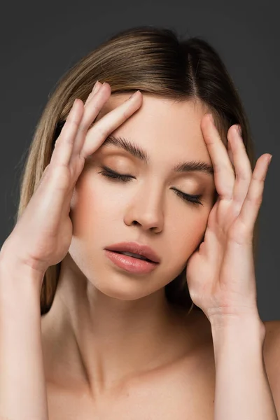 Mujer joven con los ojos cerrados y maquillaje natural tocando la cara aislada en gris - foto de stock