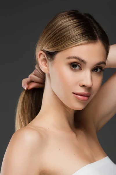 Pretty young woman with perfect skin holding hair while looking at camera isolated on grey — Stock Photo