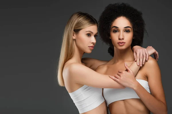 Pretty woman in white top embracing african american woman with perfect skin isolated on grey — Stock Photo