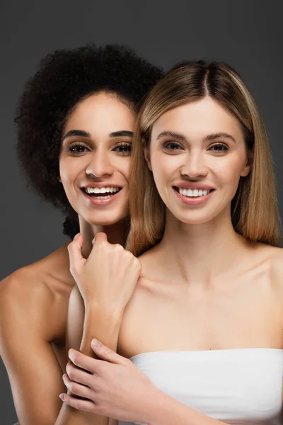 Jeunes et heureuses femmes multiethniques aux épaules nues souriant à la caméra isolée sur gris — Photo de stock