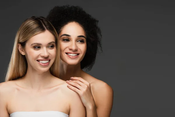 Happy multiethnic women with bare shoulders and perfect skin looking away isolated on grey — Stock Photo