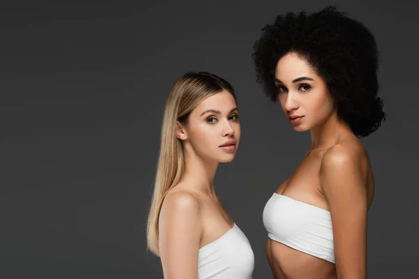 Young multiethnic women in white tops and natural makeup looking at camera isolated on grey — Stock Photo