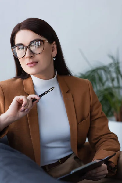 Brunette psychologist in eyeglasses looking at camera while holding clipboard and pen — Stock Photo
