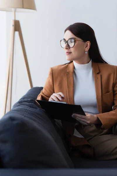 Psychologue en tenue formelle et lunettes regardant loin tout en étant assis sur le canapé avec presse-papiers — Photo de stock