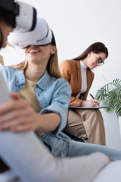 Smiling psychologist writing on clipboard near blurred interracial same sex couple gaming in vr headsets — Stock Photo