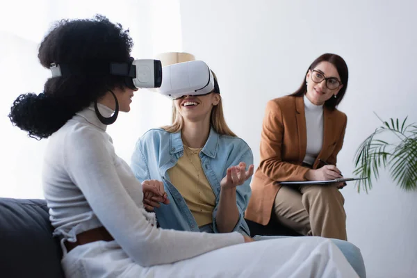 Happy interracial lesbian couple in vr headsets holding hands near blurred psychologist writing on clipboard — Stock Photo