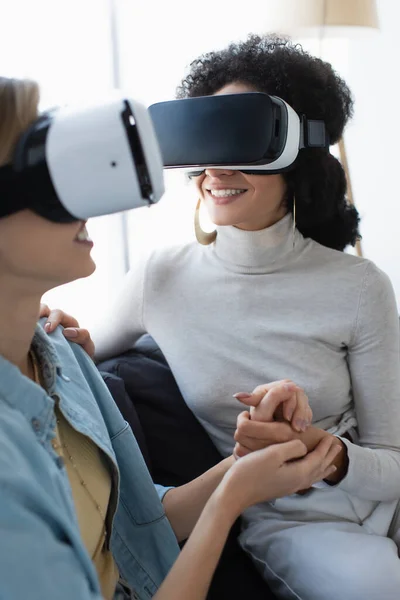 Young and happy multiethnic lesbian women gaming in vr headsets while holding hands at home — Stock Photo