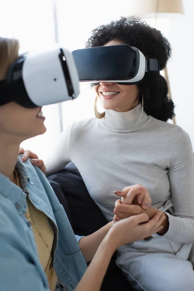 Mujeres lesbianas multiétnicas jóvenes y felices jugando en auriculares vr mientras se toman de la mano en casa - foto de stock