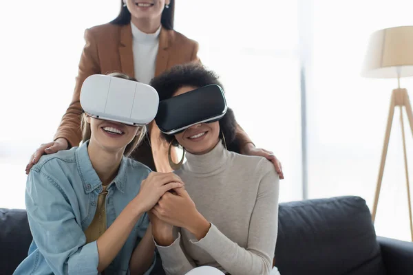 Sorrindo casal lésbico interracial em fones de ouvido vr de mãos dadas perto psicólogo turvo — Fotografia de Stock