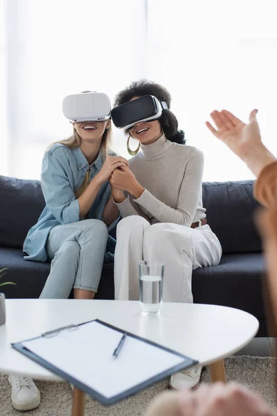 Smiling multiethnic lesbian couple in vr headsets holding hands near blurred psychologist pointing with hand — Stock Photo