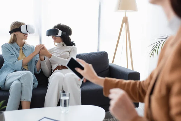 Smiling same sex couple in vr headsets holding hands on sofa near blurred psychologist with smartphone — Stock Photo