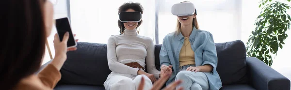 Interracial lesbian couple in vr headsets smiling near blurred psychologist with smartphone, banner — Stock Photo