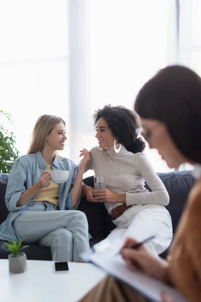 Blurred psychologist writing near multiethnic lesbian couple talking on couch — Stock Photo
