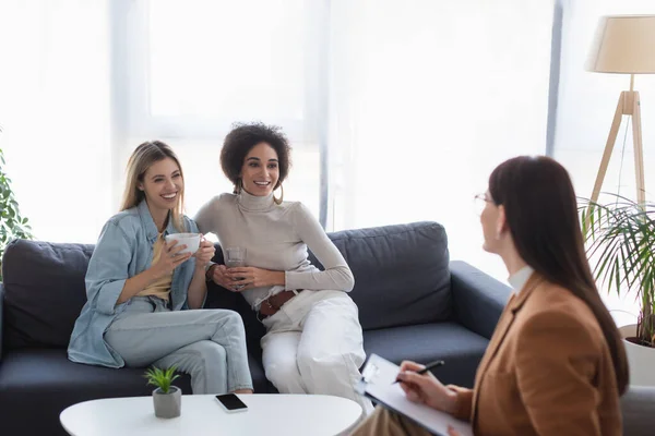 Blurred psychologist writing on clipboard near happy interracial couple of lesbians — Stock Photo