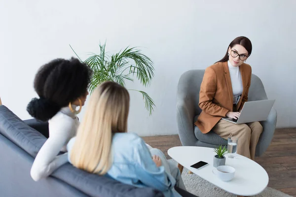 Psychologist typing on laptop while looking at interracial lesbian couple sitting on sofa — Stock Photo