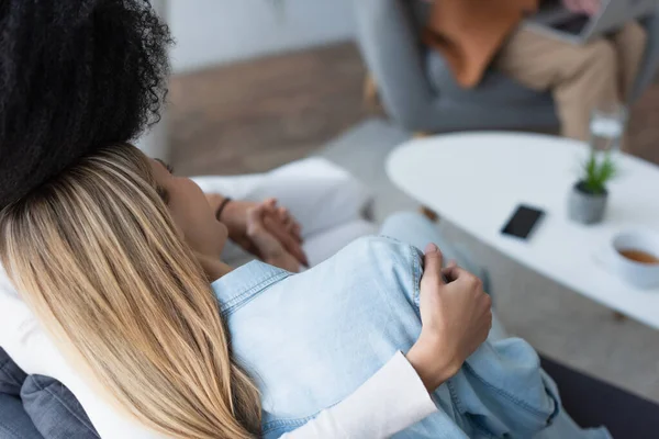 Vista de ángulo alto de lesbianas interracial abrazando durante la cita con psicólogo borroso - foto de stock