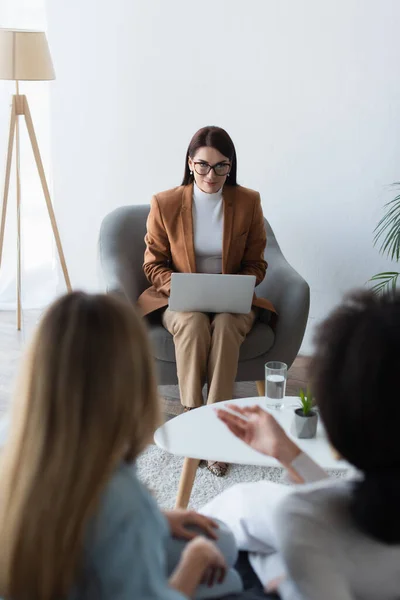 Psychologist with laptop listening to blurred interracial lesbian couple during appointment — Stock Photo