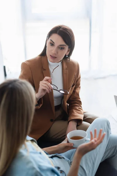 Psychologist holding eyeglasses while talking to blurred woman with cup of tea — Stock Photo