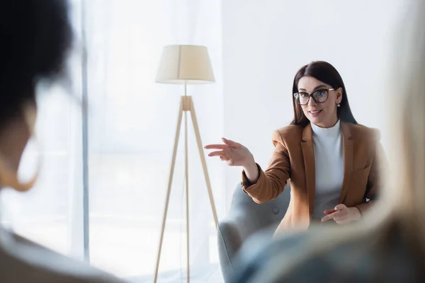 Psychologist in eyeglasses pointing with hand while talking to blurred same sex couple — Stock Photo