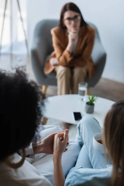 Blurred interracial same sex couple holding hands during psychological consultation — Stock Photo