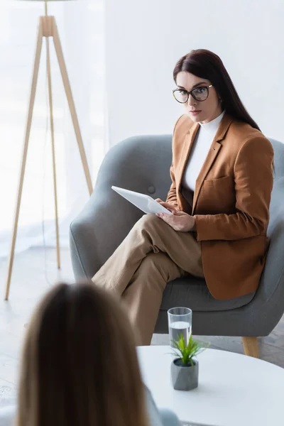 Psychologist in eyeglasses using digital tablet during consultation with blurred woman — Stock Photo