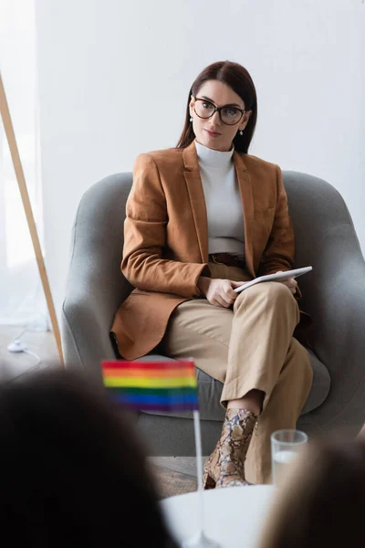 Psychologist with digital tablet sitting near patient and lgbt flag on blurred foreground — Stock Photo