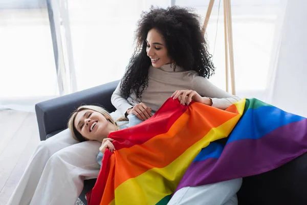 Cheerful multiethnic lesbian women with lgbt flag on couch at home — Stock Photo