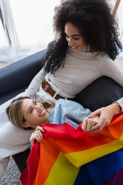 Feliz interracial lésbicas mulheres olhando umas para as outras no sofá perto lgbt bandeira — Fotografia de Stock