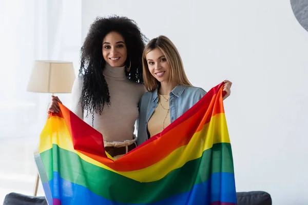 Jovens e alegres lésbicas multiétnicas olhando para a câmera enquanto de pé com bandeira lgbt — Fotografia de Stock