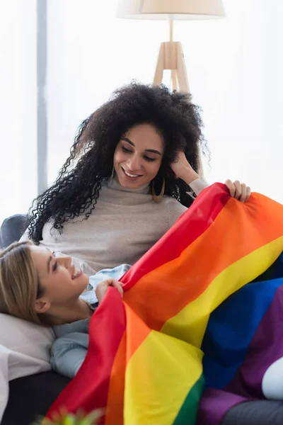 Happy lesbian woman lying on couch with lgbt flag near african american girlfriend — Stock Photo