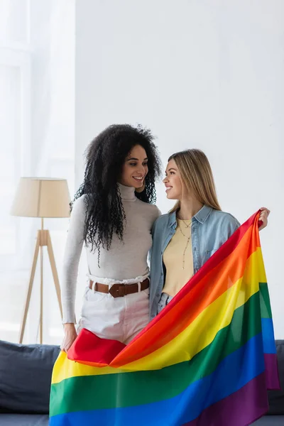 Jóvenes lesbianas multiétnicas mujeres sonriendo el uno al otro mientras sostiene la bandera lgbt - foto de stock